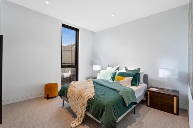 A bright, contemporary bedroom with a neatly made bed dressed in deep green and pale teal bedding, layered cushions, and a textured cream throw. The room has light grey walls, soft carpet flooring, minimalist white bedside lamps, a dark timber bedside table, a small mustard ottoman, and a tall black-framed window.