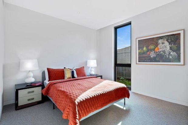 Bright, compact contemporary bedroom with a low-profile bed dressed in a red quilted coverlet, matching red and patterned cushions, white bedside tables and lamps, grey carpet, white walls, a tall narrow black-framed window, and framed artwork.