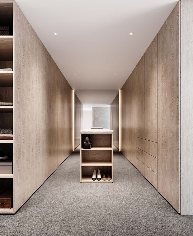 A refined walk-in wardrobe with full-height light timber cabinetry lining both sides of a narrow room. Integrated drawers and open shelving provide storage, while a central open shelving island displays shoes and accessories. The space has grey carpet flooring, a white ceiling with recessed downlights, and softly illuminated timber-lined niches at the far end.