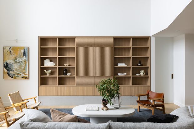 A refined contemporary living room centered on a large built-in timber shelving and media wall with open cubbies and a vertically slatted feature panel. The room includes a pale stone or composite oval coffee table, textured neutral sectional sofa, mid-century-inspired wooden lounge chairs, a large abstract artwork, and understated decorative objects. White walls, timber flooring, and abundant natural light create a warm, calm interior.
