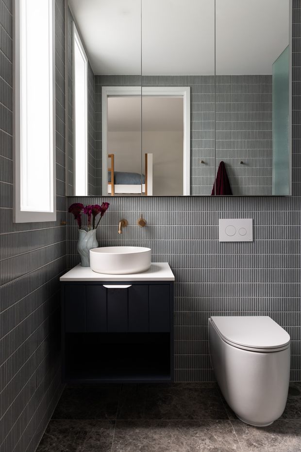 Compact contemporary bathroom featuring dark grey vertically ribbed wall tiles, dark stone-look floor tiles, a floating black vanity with a white vessel basin, wall-mounted tapware, a frameless mirrored cabinet, and a modern white toilet. A bedroom is visible through the doorway in the mirror reflection.