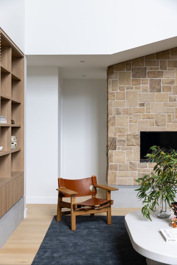 A refined contemporary living room with a built-in timber shelving unit, pale timber flooring, a sculptural leather-and-wood lounge chair, dark rug, rounded white coffee table, and a substantial natural stone fireplace wall. The room has a clean white ceiling and walls with recessed lighting, creating a warm, minimalist atmosphere.