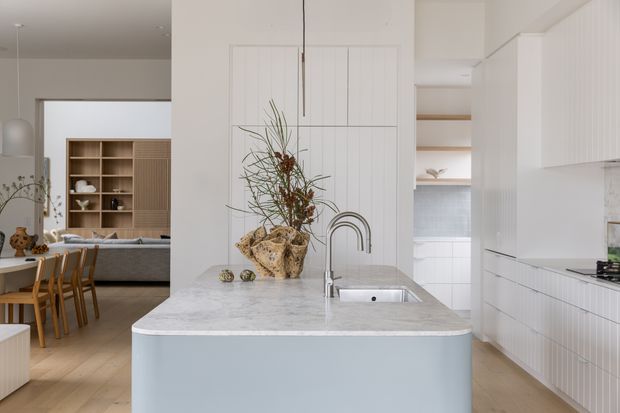 Bright open-plan kitchen with a large pale stone island, integrated sink and curved brushed-metal tap. White vertical-panel cabinetry and matching wall panelling create a clean, refined backdrop, while light timber flooring and an adjacent dining and living area add warmth. Open shelving and decorative styling are visible beyond the kitchen.