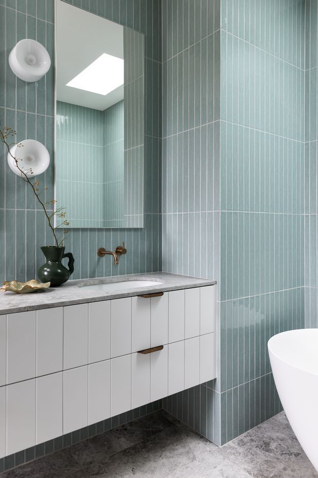 A refined contemporary bathroom with floor-to-ceiling pale blue-green vertically laid wall tiles, a floating white vanity with slim integrated drawers and bronze pulls, a grey stone benchtop with inset basin and wall-mounted bronze tapware, and a partially visible freestanding white bathtub. A large frameless mirror, sculptural wall lights, dark stone-look flooring, and minimal decorative styling complete the space.