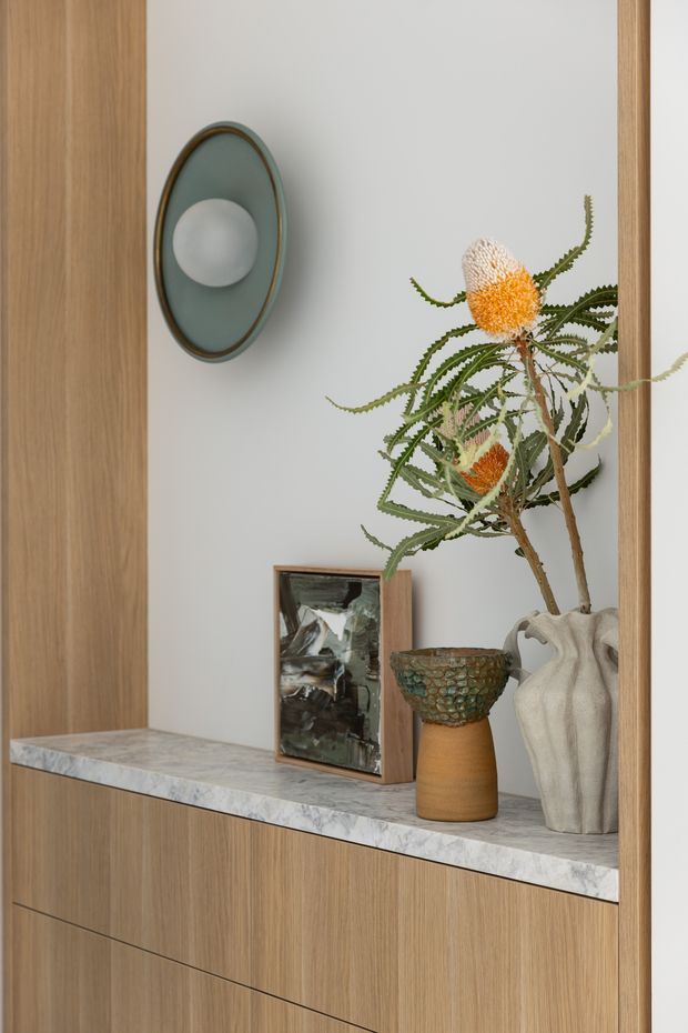 Close-up of a refined built-in timber display cabinet or entryway joinery. The warm wood cabinetry surrounds a pale marble shelf, styled with sculptural ceramics, a framed artwork, dried banksia flowers, and a round wall light with a muted green shade and bronze rim.