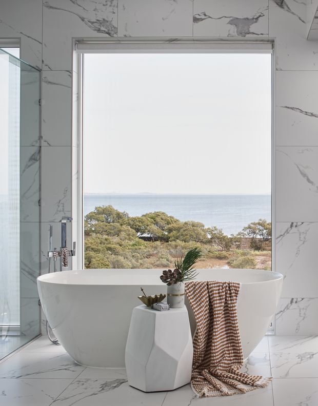 A bright, luxurious bathroom featuring a freestanding white bathtub positioned in front of a large picture window overlooking coastal vegetation and the sea. The walls and floor are clad in white marble-look tiles with prominent grey veining, and a glass shower enclosure is visible at the left. A sculptural white side table, potted plant, and textured brown-and-white towel add styling details.