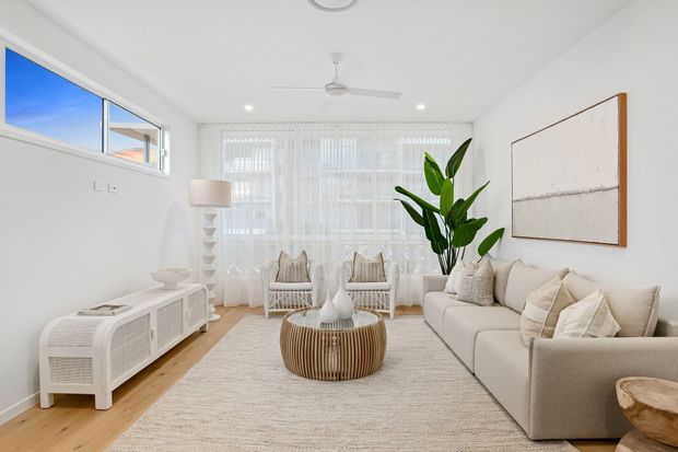 Bright, airy living room with white walls, sheer floor-to-ceiling curtains, a large neutral sectional sofa, woven round coffee table, white media console, rattan lounge chairs, indoor tropical plant, and abstract wall artwork. The room has abundant natural light and a relaxed coastal-inspired palette.
