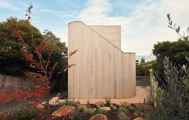 A sculptural, compact building exterior clad in vertically ribbed pale timber, with a rounded corner and stepped, angular roofline. It sits within a landscaped garden of shrubs, grasses and stone edging, with a metal railing and glimpses of a blue water or sky horizon beyond. The image has a warm, natural contemporary architectural character.