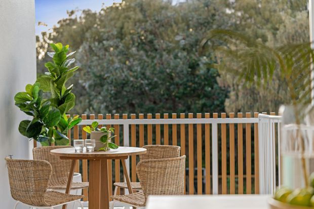 A bright, inviting outdoor dining terrace with a round timber table, woven wicker dining chairs, potted greenery, and a vertical timber-slatted balustrade. The terrace overlooks leafy vegetation and is framed by light-coloured walls and glazing.