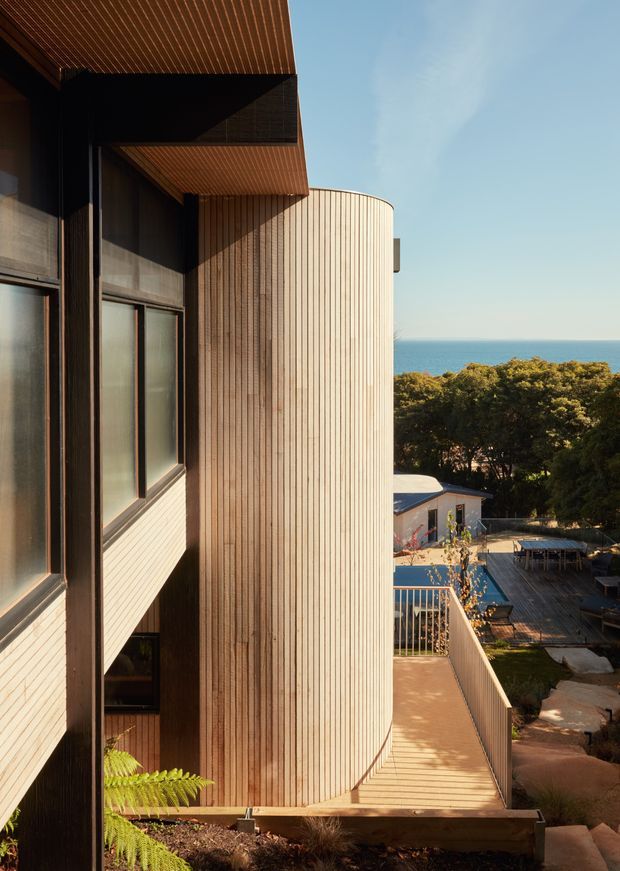 Exterior architectural view of a contemporary coastal residence, focused on a tall curved volume clad in closely spaced vertical timber battens. Dark-framed glazing and projecting timber soffits appear on the left, while a raised timber deck and balustrade lead toward a lower outdoor entertaining area. Trees and the ocean are visible beyond the property.