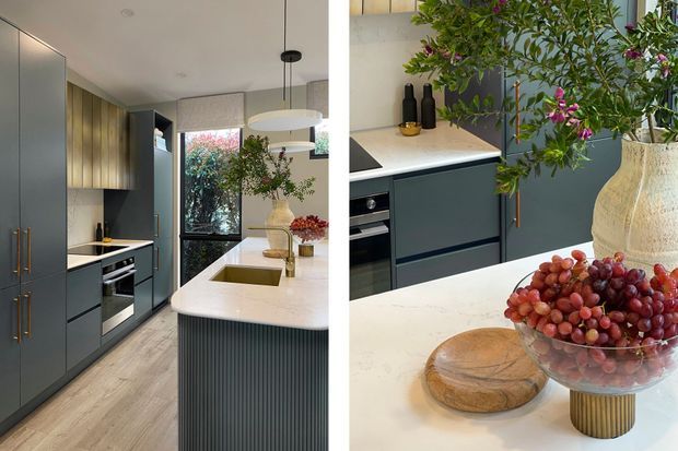 A contemporary kitchen shown in a split view, featuring dark blue-grey full-height cabinetry with slim brass-toned handles, a light stone island benchtop with integrated double sinks, fluted vertical detailing on the island, warm timber cabinetry, integrated appliances, and pendant lighting. The close-up view highlights the pale stone surfaces, dark cabinetry, brass hardware, and styled countertop.