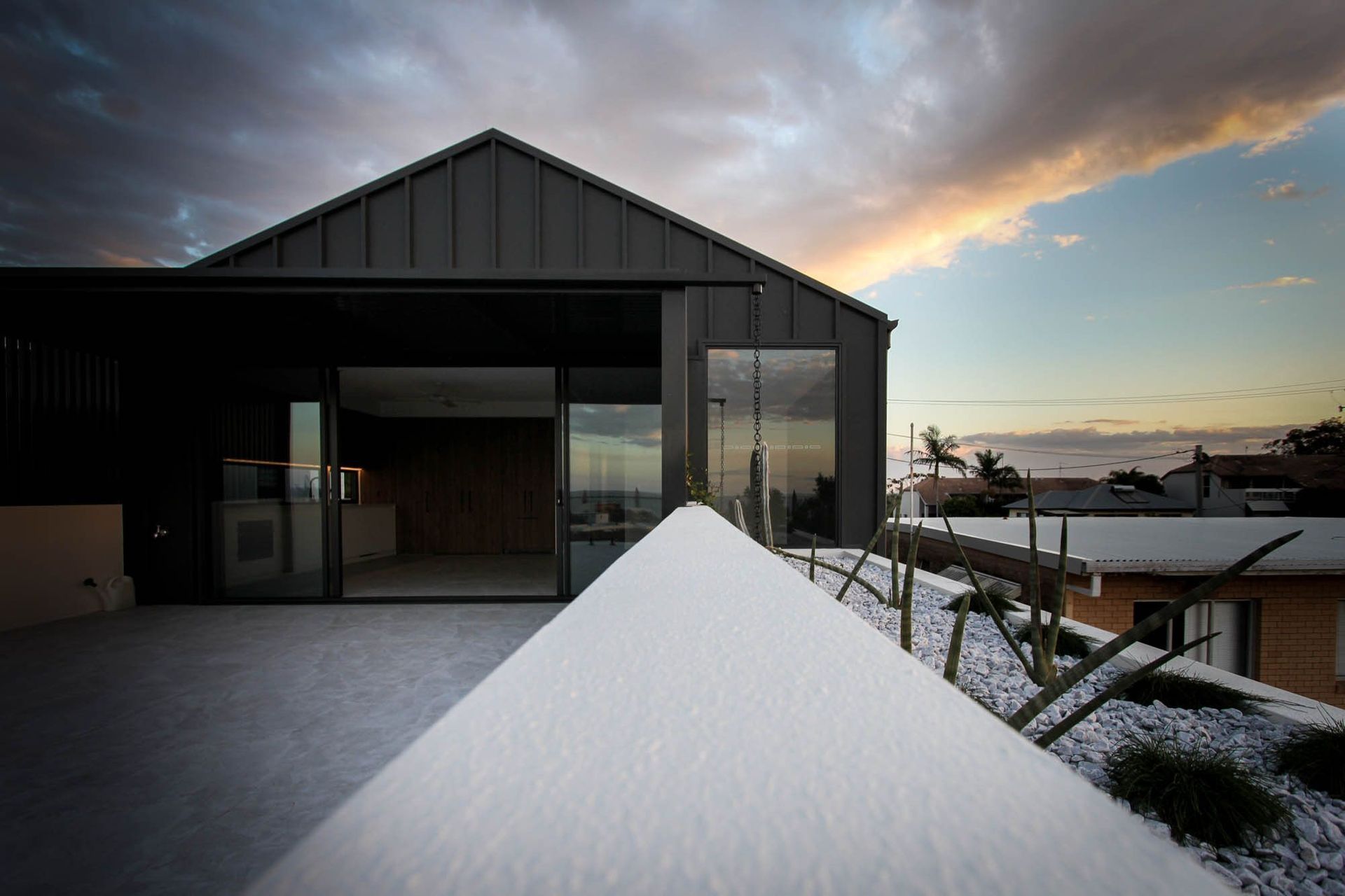 Anderson Residence - Exterior. Dark gabled form opening to a terrace with coastal views at sunset.