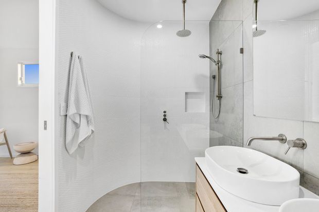 Bright contemporary bathroom with a curved, glass-enclosed walk-in shower, dual overhead rain showerheads, wall-mounted hand shower, recessed niche, and a white vessel basin on light timber cabinetry. White textured wall tiles, pale grey floor tiles, and brushed metal fittings create a clean, minimalist appearance.