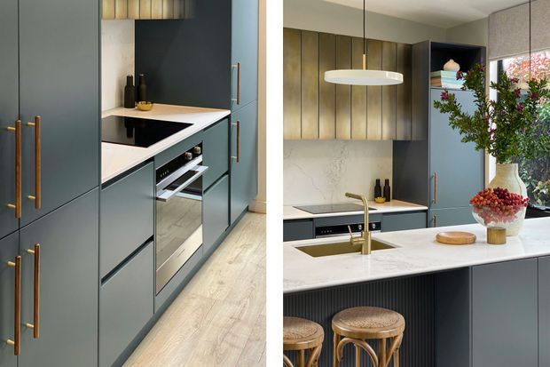 Contemporary kitchen featuring tall matte blue-grey cabinetry with slim brass pulls, warm vertical timber wall panelling, integrated appliances, a pale stone island benchtop, brass tapware, and woven timber bar stools. The space is softly lit and styled with restrained decorative objects and greenery.