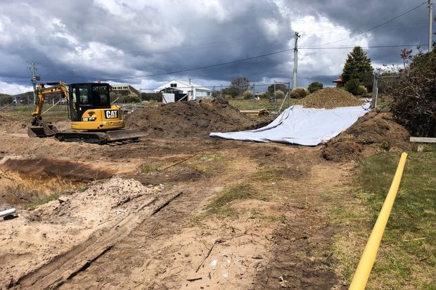 A residential construction site showing a yellow excavator, extensive earthworks, exposed soil, a white ground cover or geotextile sheet, utility poles, and partially visible suburban buildings under a cloudy sky.
