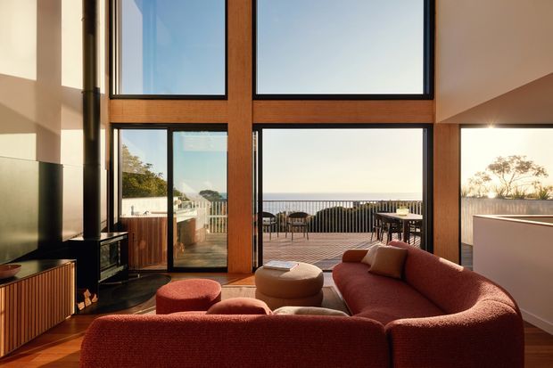 A warm, contemporary living room with a large curved rust-coloured sectional sofa and coordinating ottomans, centred around a dark fireplace. Floor-to-ceiling timber-framed glazing opens the room visually to a timber outdoor deck and expansive coastal or waterfront-like horizon. The room has a high ceiling, strong natural light, and a refined indoor-outdoor connection.