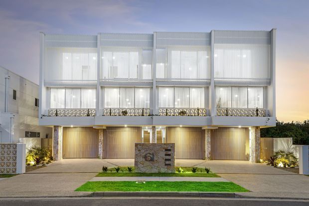 Front exterior view of a contemporary multi-unit residential building at dusk, featuring three vertically articulated dwellings with white vertical screens, large glazed upper-level openings, recessed balconies with decorative patterned balustrades, ground-level garages, warm facade lighting, and a landscaped central entry feature.