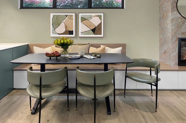 A stylish dining area with a long dark dining table and four olive-green upholstered chairs, set in front of a built-in banquette with cushions. The room features warm timber-look flooring, pale green walls, low horizontal windows, abstract framed artwork, integrated cabinetry, and a textured stone fireplace wall with a partially visible firebox. The overall scheme is refined, warm, and contemporary with mid-century influences.