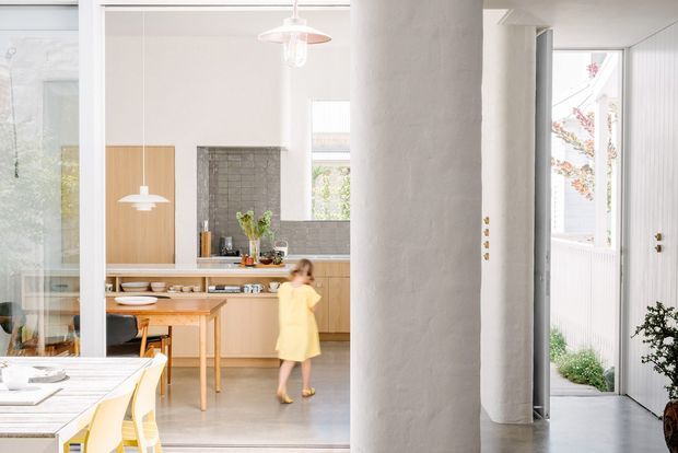 Bright contemporary open-plan interior with a dining area in the foreground and a warm timber kitchen beyond. White plastered structural columns frame the space, with pale cabinetry, a timber dining table, pendant lights, large glazed openings, and a small planted entry courtyard visible at right.