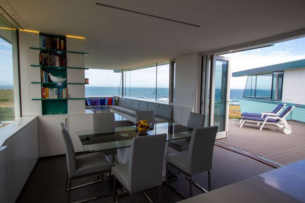 Open-plan contemporary dining and living area overlooking the ocean, with floor-to-ceiling glazing, a glass dining table, upholstered dining chairs, an L-shaped sectional sofa, and built-in shelving. Large sliding glass doors open onto a timber deck with poolside loungers and a small turquoise-clad structure beyond.