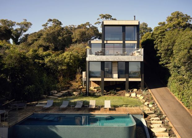 A narrow, multi-storey contemporary house set among dense bushland, viewed from the rear. The building has a strongly vertical composition with dark-framed glazing, pale cladding, a flat roof, and elevated lower levels supported by slender posts. A large balcony overlooks a landscaped lawn and swimming pool, with a paved driveway rising along the right side.