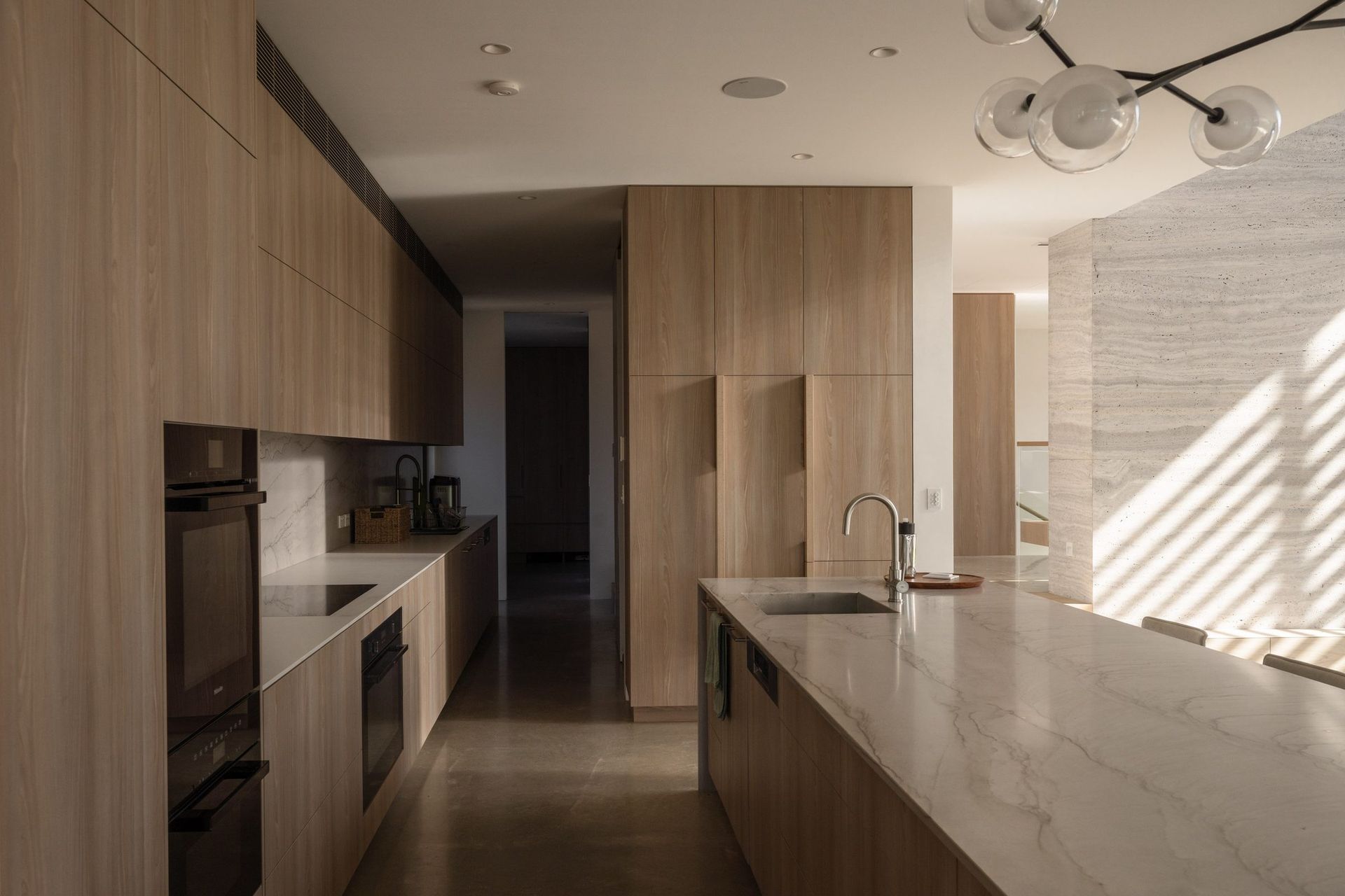 Decade House - Kitchen. Long kitchen with timber joinery, integrated appliances and a stone island.