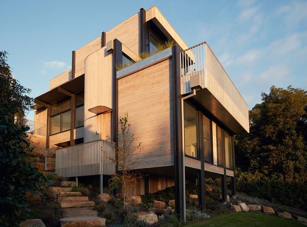 Exterior view of a contemporary multi-storey house set on a landscaped, sloping site. The building features extensive warm timber cladding, dark slender structural posts and window frames, projecting balconies, vertical metal balustrades, large glazed openings, and a strongly articulated angular form. Rock landscaping and mature trees surround the elevated house.