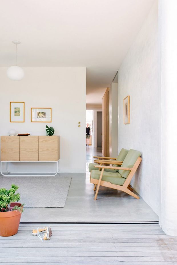 Bright minimalist entry and living area with white walls, pale grey polished concrete flooring, and a light timber deck transition. A light oak sideboard with slim metal legs sits beneath framed artwork, while a green upholstered timber lounge chair occupies the right side. A hallway extends toward the rear, and potted plants and shoes appear near the threshold.