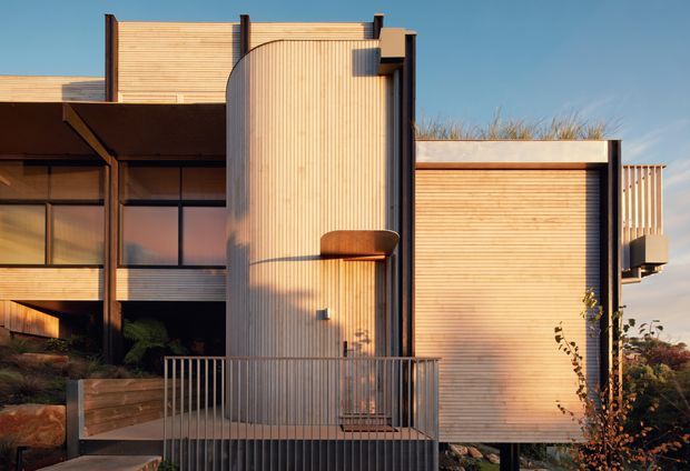 A close exterior view of a contemporary residential building composed of warm timber-clad volumes, dark structural framing, large glazed openings, and a prominent curved vertical timber-clad facade element. A metal-railed terrace and landscaped foreground sit at the base, with low evening sunlight emphasizing the building's layered geometry and texture.