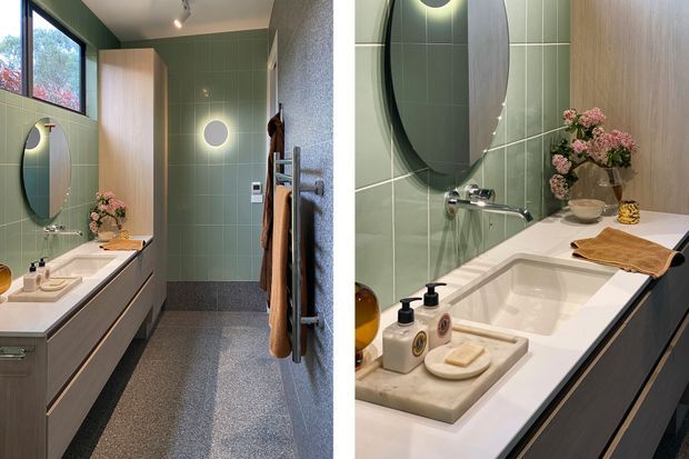 A contemporary bathroom featuring a long floating vanity with an integrated white basin, pale green vertical wall tiles, a large oval mirror, timber cabinetry, terrazzo-style flooring, and a freestanding towel rail. Natural light enters through a high window, while warm integrated lighting and floral styling add softness.