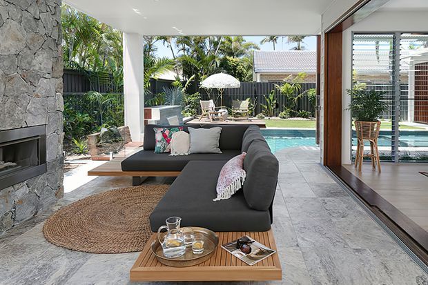 An airy, open-plan outdoor living area beside a swimming pool, featuring a large dark sectional sofa, woven round rug, timber coffee table, and a substantial rough-stone fireplace wall. Full-height glazed sliding doors connect the covered lounge to the interior, with tropical planting and a fenced garden visible beyond.