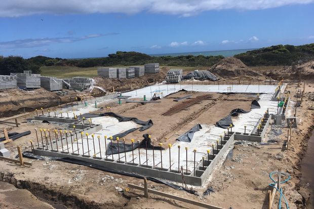 A construction-site view showing a newly poured concrete slab and perimeter footings with exposed reinforcing steel, formwork, plumbing conduits, drainage elements, and construction materials. The site is set in a sandy coastal landscape with low scrub and ocean visible in the background.