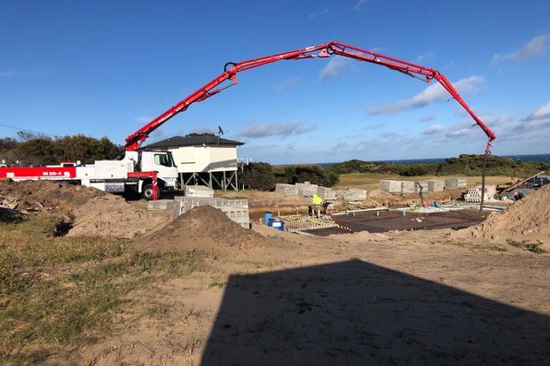 A construction site showing a large red concrete pump truck extending its boom over a partially built concrete foundation. Excavated earth, concrete block walls, reinforcing materials, and workers are visible, with a coastal horizon in the background.