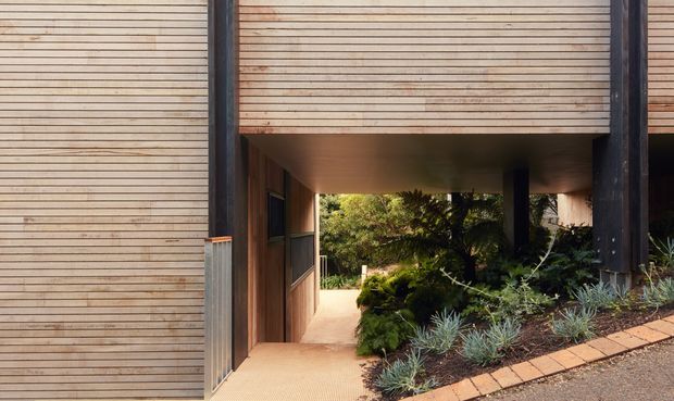 Exterior architectural view of a contemporary building with a covered passageway or entry recess. The facade features finely horizontal-lined timber cladding, dark vertical structural posts, and a warm-toned paved walkway bordered by low drought-tolerant landscaping. The image looks through the sheltered opening toward a planted courtyard or garden.