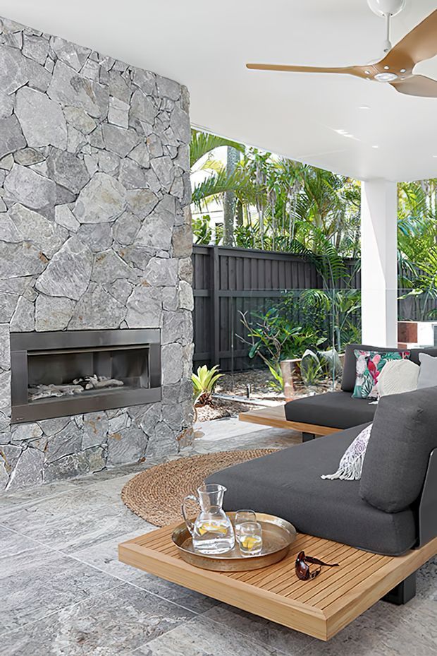 Bright, open-sided outdoor living area with a large irregular grey stone feature wall and built-in fireplace. The space has dark charcoal upholstered seating, a timber slatted platform and coffee table, a woven round rug, a ceiling fan, and lush tropical planting behind a dark privacy fence. Grey stone paving continues throughout the covered entertaining area.
