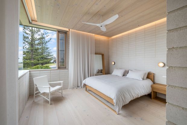 Bright, serene contemporary bedroom with a low-profile timber bed, white bedding, pale timber flooring and ceiling, and a white horizontally panelled feature wall. Full-height sheer curtains frame a large window overlooking trees and water, while integrated warm cove lighting and a white ceiling fan add comfort. A white armchair sits beside the window, and a small timber bedside table and wall sconces complete the room.