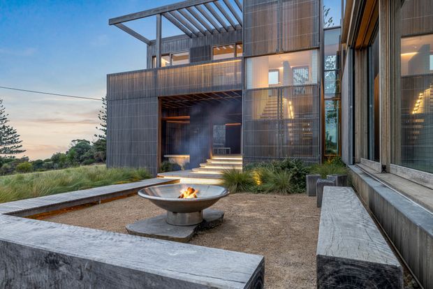 Contemporary multi-level residential exterior at dusk, featuring dark vertical cladding, extensive glazing, a pergola-like roof structure, recessed entry steps, and a landscaped courtyard with a central fire bowl and built-in concrete seating.