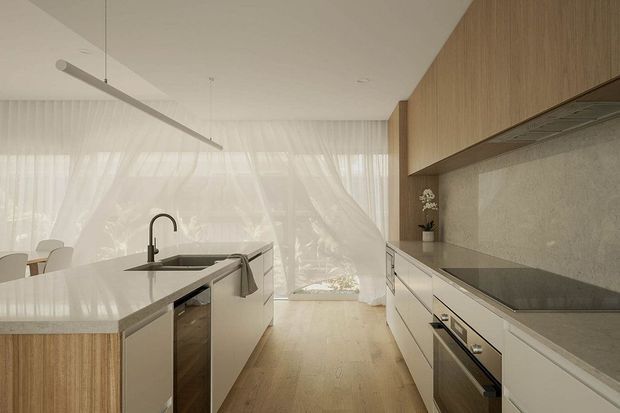 A bright, refined contemporary kitchen viewed along a central walkway. The room features a large light stone island with an integrated sink and black tap, pale cabinetry with timber drawer fronts, a full-height textured stone splashback, timber overhead cabinetry, a dark cooktop and built-in ovens. Sheer curtains span the glazed rear wall, and a slim linear pendant light hangs above the island. A glimpse of the dining area is visible at left.