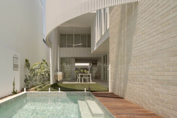 A bright contemporary courtyard viewed from beside a small swimming pool. The space features a pale rendered wall, curved white architectural canopy and upper-level screening, extensive glazing, a light brick feature wall, timber decking, landscaped planting, and a double-height glazed living area beyond the courtyard.
