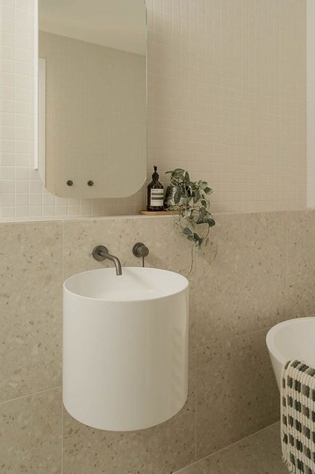 A minimalist bathroom vanity featuring a cylindrical white vessel basin with wall-mounted brushed metal tapware, a large rounded rectangular mirror, pale small-format wall tiles, and beige-grey stone-look wall and floor tiles. A small plant and soap dispenser sit on the ledge, with part of a freestanding bathtub visible at right.