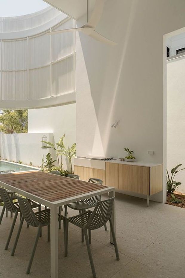 A bright, modern covered outdoor dining area beside a pool, featuring a large timber dining table with woven chairs, a minimalist built-in outdoor kitchen or serving cabinet in light timber, pale concrete flooring, lush planting, and a dramatic curved upper-level screened structure with extensive vertical glazing or screening. The space has a clean, airy indoor-outdoor character.
