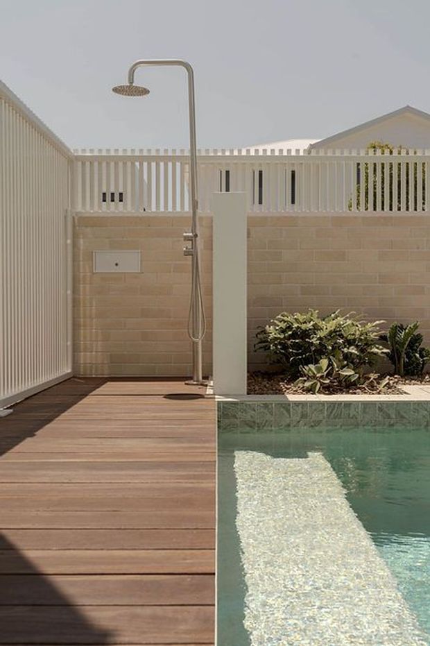 A compact outdoor pool courtyard with a timber deck, narrow rectangular swimming pool, integrated pale stone or concrete-block privacy wall, and a freestanding outdoor shower. White vertical slatted fencing and a raised garden bed with greenery enclose the space. The palette is warm timber, sandy beige masonry, white fencing, and muted aqua pool water in bright daylight.