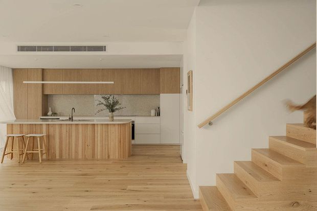 Warm, minimalist open-plan kitchen and stairway interior with light timber flooring, a timber island and cabinetry, pale stone benchtops, and white walls. A stair with timber treads and a simple timber handrail rises along the right side, while integrated cabinetry and concealed appliances create a clean, contemporary appearance.