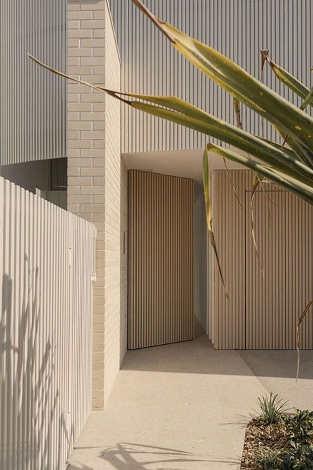 A minimalist residential entry courtyard with a pale brick wall, vertically ribbed metal or timber-look cladding, a tall slatted entry gate, and a pale textured concrete path. Strong linear shadows and sculptural drought-tolerant planting add visual interest to the warm, neutral facade.