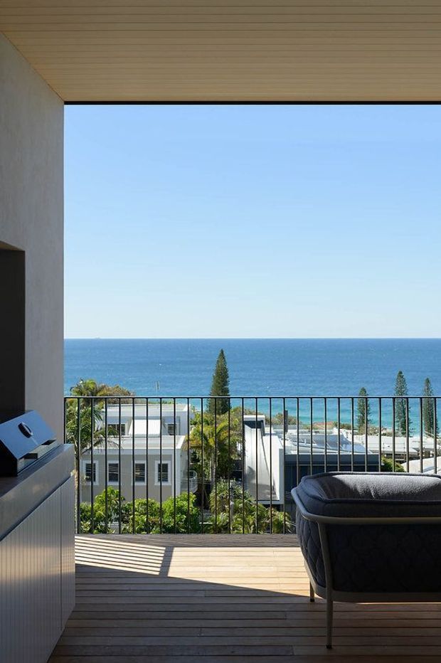 A contemporary open-plan living area opens onto a timber-decked balcony with black metal balustrading and expansive ocean views. The space features a pale timber-lined ceiling, light rendered walls, built-in cabinetry, and dark upholstered seating. Modern residential buildings and coastal vegetation are visible beyond the balcony.