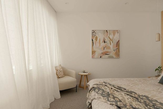 A softly lit, neutral-toned bedroom with sheer floor-to-ceiling curtains, a cream upholstered armchair, a small round timber side table, and a bed dressed in layered beige and patterned bedding. Abstract artwork and a simple pendant light add restrained decorative accents.