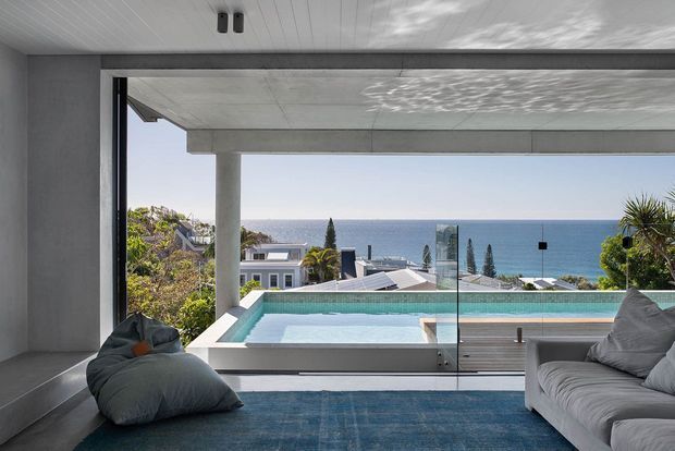 A contemporary living room opens onto a balcony or terrace with a glass balustrade and a long rectangular swimming pool. Floor-to-ceiling glazing frames a clear ocean view, while a grey sofa, beanbag chair, and blue rug furnish the calm, minimalist interior. The room has a white slatted ceiling, substantial concrete framing, and a strong indoor–outdoor connection.