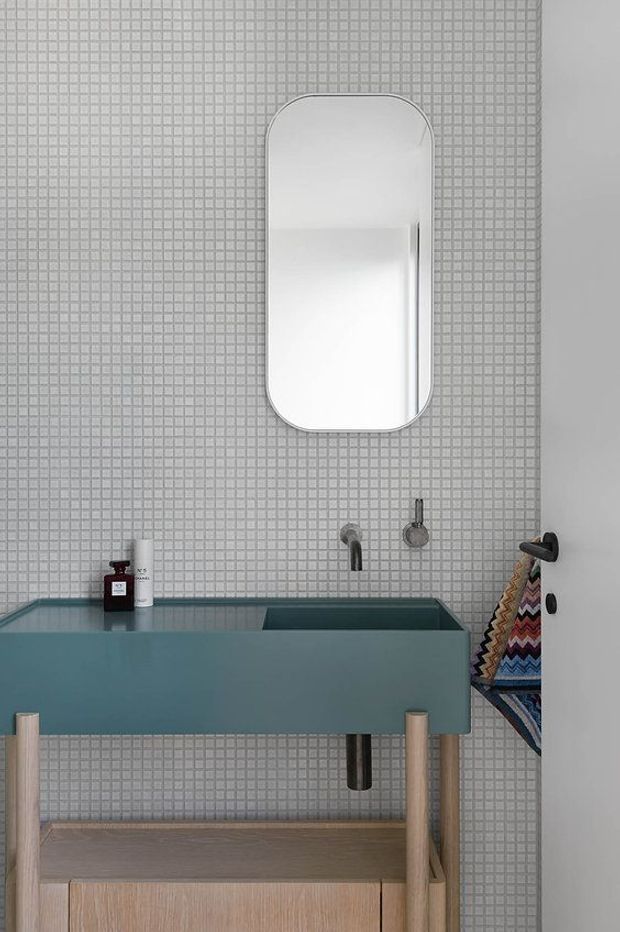 A close-up view of a contemporary bathroom vanity. The wall is clad in small square light-grey mosaic tiles, with a rounded-rectangle frameless mirror above a teal integrated basin and wall-mounted chrome tapware. A timber-framed open shelf and cabinetry sit beneath the vanity, with a colourful patterned towel hanging at the side.