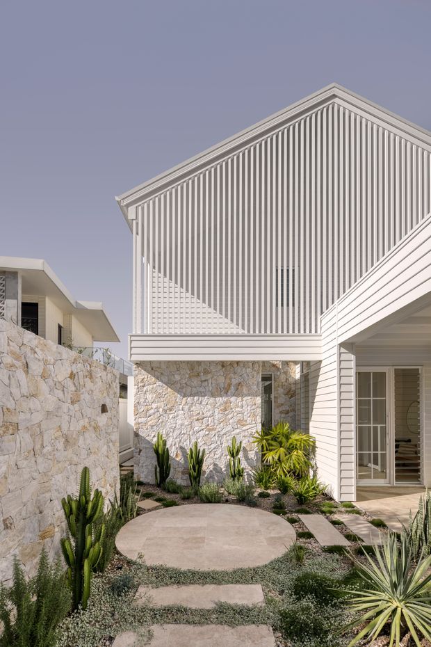 Exterior view of a contemporary residential courtyard or entry garden. White weatherboard-clad buildings with steep gabled roof forms and a striking vertical slatted gable screen surround a landscaped path. Rough pale natural-stone walls, concrete stepping slabs, gravel, and drought-tolerant cacti and succulents create a modern desert-style courtyard.