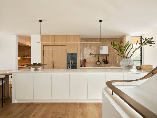 A warm, refined contemporary kitchen viewed from an adjacent stair or landing. The room features a large white island with integrated cabinetry and a pale benchtop, timber flooring, light timber cabinetry, a textured timber-toned splashback or feature wall, integrated appliances, and minimal black pendant lighting. A curved white stair with timber handrail occupies the foreground, while plants and simple kitchen accessories add natural detail.